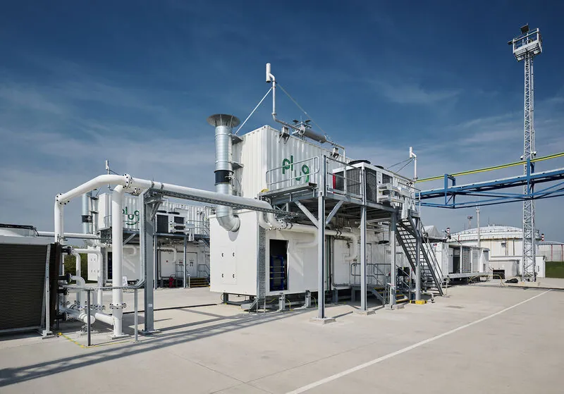 Photo of the Plug Power electrolyser: containerised modules bearing the brand logo, with pipework, stairs and steel walkways at an industrial site
