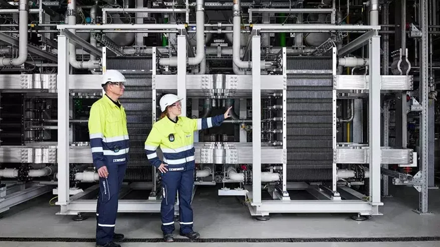 Photo of two technicians in hard hats and high-visibility clothing in front of a row of PEM electrolyser stacks inside a hall