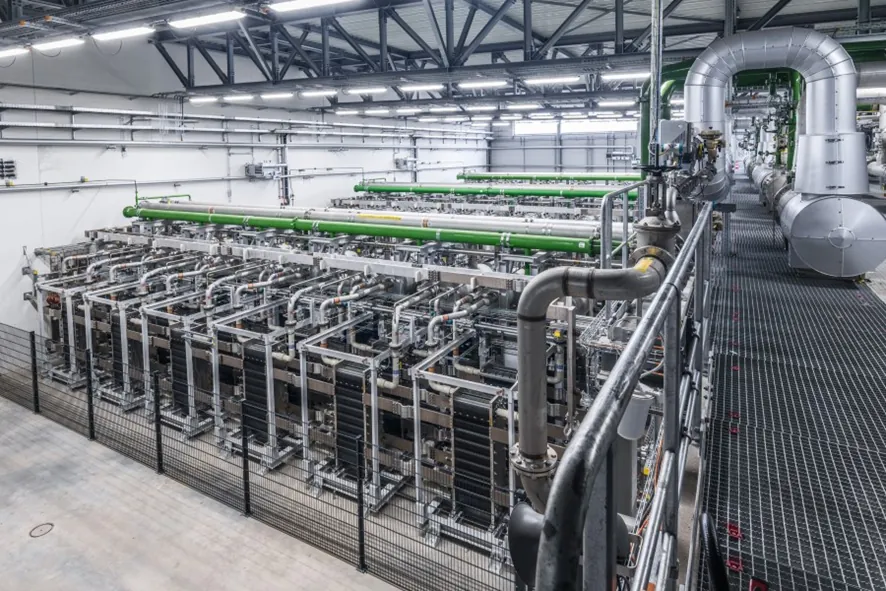Fotografía de una gran nave de electrólisis con filas de stacks, colectores verdes y plateados y una pasarela elevada